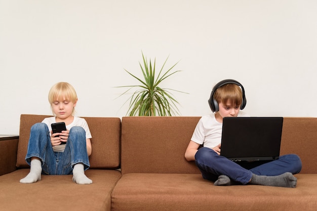 Zwei Kinder sitzen auf der Couch mit Gadgets Smartphone und Laptop. Generation Alpha.