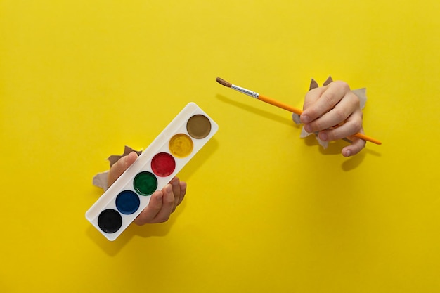 Zwei Kinderhände, die durch ein zerrissenes gelbes Papierblatt gestreckt sind, halten Pinsel und Farben zum Zeichnen