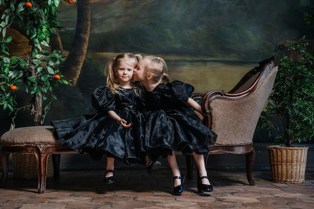 Zwei kleine italienische Mädchen sitzen auf einer alten Couch in schwarzen Kleidern im Studio mit Pflanzen