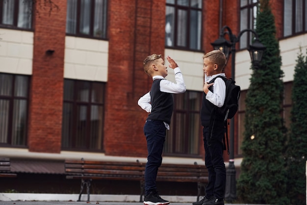 Zwei kleine jungen in schuluniform, die im freien ist, geben zusammen high five in der nähe des bildungsgebäudes.