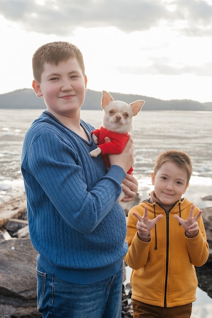 Zwei kleine Jungen stehen in den Armen eines Chihuahua-Hundes am Fluss. Das Konzept der Freundschaft und Brüderlichkeit. Erholung im Freien.