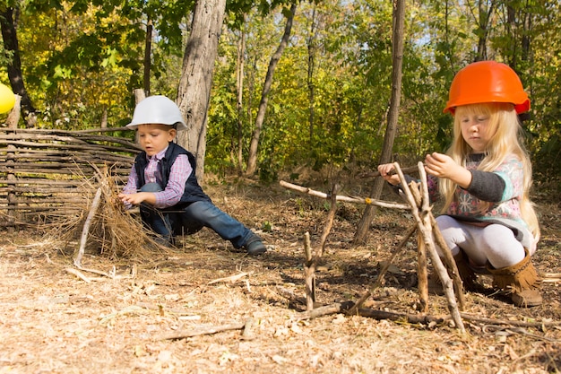 Zwei kleine Kinder, die vorgeben, Bauarbeiter oder Bauarbeiter zu sein, die Schutzhelme tragen und Rahmen aus Zweigen und Ästen im Freien im Wald schaffen