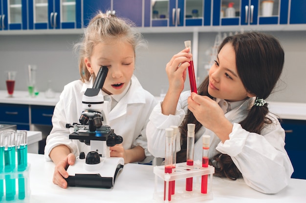 Zwei kleine Kinder im Laborkittel lernen Chemie im Schullabor. Junge Wissenschaftler in Schutzbrillen machen Experimente im Labor oder im Chemikalienschrank. Durch das Mikroskop schauen