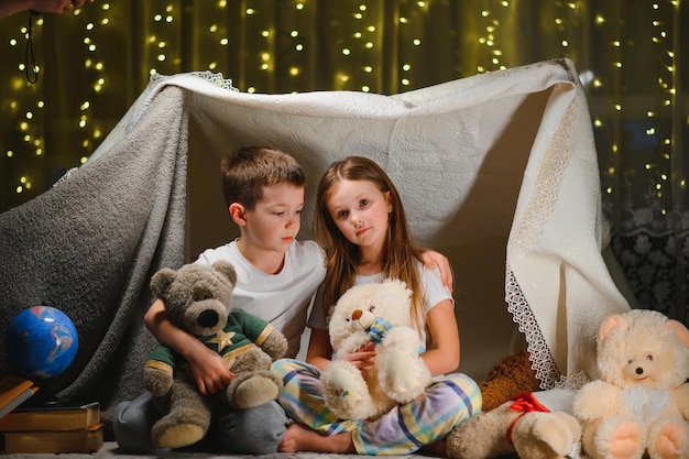Zwei kleine Kinder spielen abends zu Hause, um ein Campingzelt zu bauen, um mit einer Taschenlampe Bücher zu lesen und drinnen zu schlafen. Konzept von: Spiel, Magie, Kreativität, Alarmsysteme