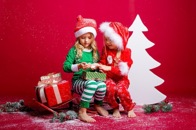 Zwei kleine Mädchen auf Weihnachtsmützen auf Rot sitzen nahe einem weißen Weihnachtsbaum mit Geschenken