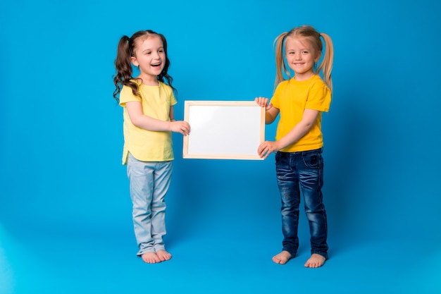 zwei kleine Mädchen, die mit einem leeren Reißbrett auf einem blauen Hintergrund lächeln