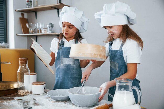 Zwei kleine Mädchen in blauer Kochuniform bereiten Essen zu, indem sie ein Sieb in der Küche verwenden und das Quittungsbuch lesen