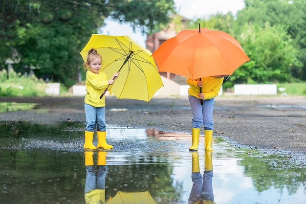 Zwei kleine Mädchen mit knallgelben und orangefarbenen Regenschirmen laufen in Gummistiefeln durch die Pfützen