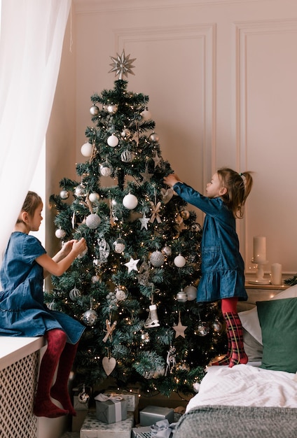 Zwei kleine Mädchen schmücken einen Weihnachtsbaum in einem Zimmer mit großem Bett