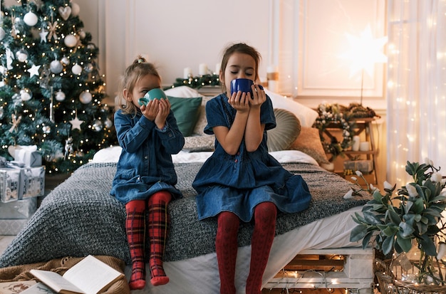 Zwei kleine Mädchen trinken Tee auf dem Bett vor dem Hintergrund eines Weihnachtsbaumes