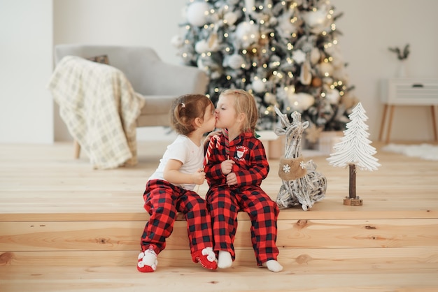 Zwei kleine Schwestern im roten Pyjama sitzen am Weihnachtsmorgen im Wohnzimmer und essen Süßigkeiten. Familienfeier, Umarmungen und Küsse