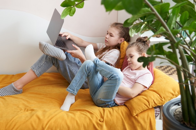 Zwei kleine Schwestern mit Laptop zu Hause auf dem Bett liegend