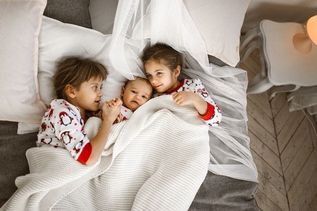 Zwei kleine Schwestern und ein kleiner Bruder liegen auf dem Bett unter der weißen Decke im hellen gemütlichen Schlafzimmer. .