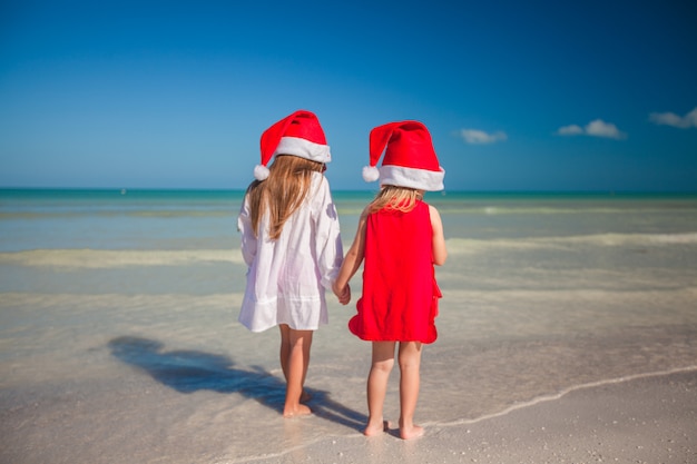 Zwei kleine süße Mädchen in Weihnachtsmützen haben Spaß am exotischen Strand