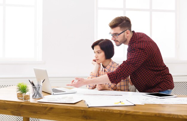 Zwei Kollegen diskutieren Architekturprojekt auf Laptop, arbeiten mit Blaupausen im modernen Arbeitsplatzinterieur, Kopierraum