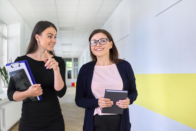 Zwei Kollegen einer Geschäftsfrau, einer Lehrerin, die auf dem Korridor des Büros, der Schule, spazieren und sprechen. Positiv lächelnde Frauen im jungen und mittleren Alter, Konzept für Büroangestellte in der Wirtschaftspädagogik