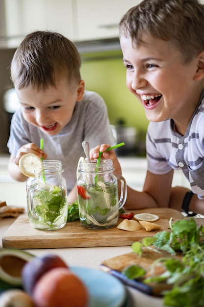Zwei lachende kleine Jungen, die erfrischenden Vitamin-Sommercocktail aus dem Glas trinken, verwenden Strohhalm und haben Spaß