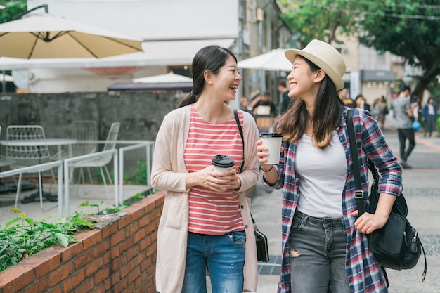 zwei lächelnde asiatische Teenagerinnen, die neben dem Garten aus rotem Backstein Einwegbecher laufen und halten. gruppe glücklicher koreanischer freundinnen entspannen sich im dorf mit pappbecher kaffee, plaudern und lachen