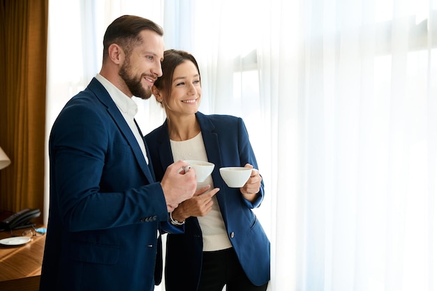 Zwei lächelnde, charmante, erfolgreiche Geschäftsleute, ein gutaussehender, selbstbewusster Mann und eine schöne Frau in Business-Anzügen, die eine Tasse Kaffee halten und selbstbewusst aus dem Fenster zur Seite schauen.