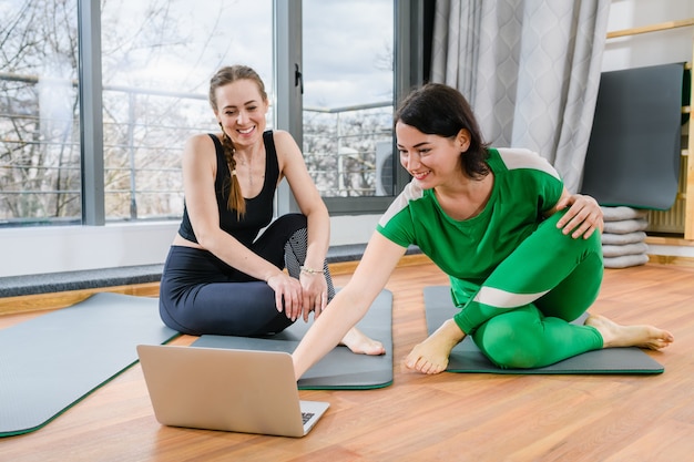 Zwei lächelnde Frauen sitzen auf Matten mit offenem Laptop-Bildschirm und bereiten sich darauf vor, mit Online-Tutorials zu trainieren