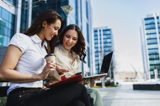 Zwei lächelnde Geschäftsfrauen, die während der Kaffeepause im Freien chatten und ein digitales Tablet verwenden.