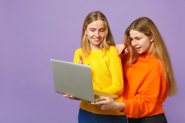 Zwei lächelnde junge blonde Zwillingsschwestern Mädchen in lebendigen bunten Kleidern halten, mit Laptop-PC isoliert auf pastellvioletter blauer Wand. Menschen-Familien-Lifestyle-Konzept.