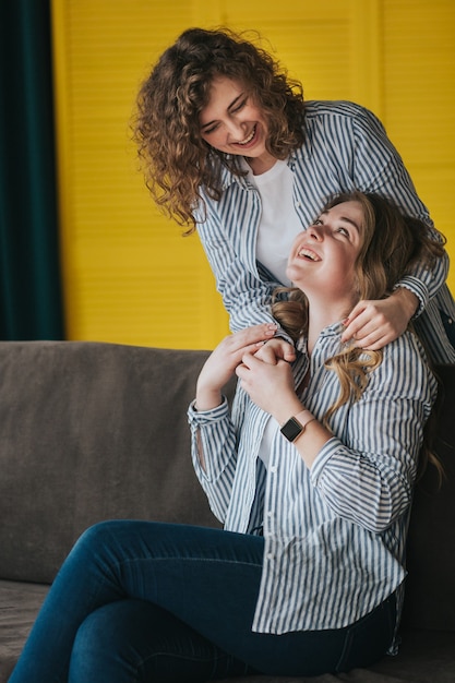 Foto zwei lächelnde junge mädchen in gestreiften hemden, jeans und turnschuhen, die auf der couch aufwerfen. studioaufnahmen.