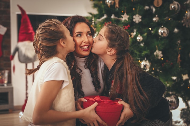 Zwei lächelnde kleine Mädchen und ihre Mutter sitzen am Weihnachtsbaum. Sie geben ihrer Mutter ein Geschenk in einer roten Schachtel und küssen sie.