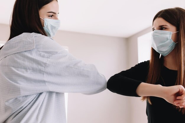 Zwei lächelnde Mädchen in schützenden Gesichtsmasken begrüßen sich auf sichere Weise mit ihrem Ellbogen in hoher Qualität
