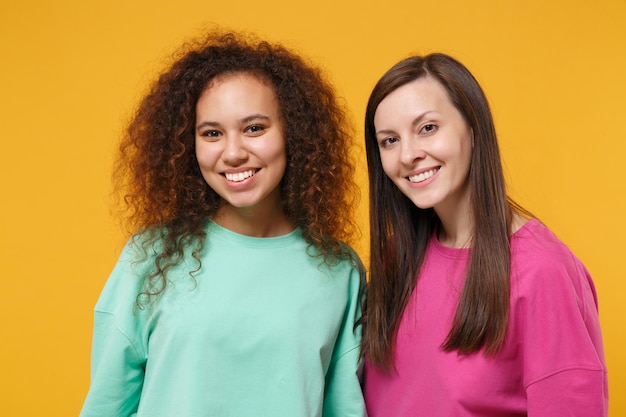 Zwei lächelnde schöne Freundinnen europäische und afroamerikanische Mädchen in rosa grüner Kleidung posieren isoliert auf gelb-orangefarbenem Hintergrund Studioporträt. People-Lifestyle-Konzept. Mock-up-Kopienbereich.