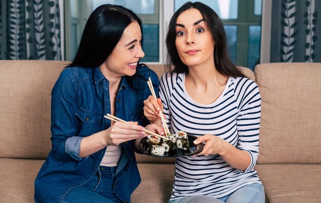 Zwei lächelnde schöne Freundinnen in moderner Freizeitkleidung sitzen auf der Couch und essen vegane Brötchen und kommunizieren