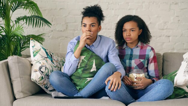 Zwei lockige Freundinnen gemischter Rassen sitzen auf der Couch und sehen sich zu Hause einen sehr gruseligen Film im Fernsehen an