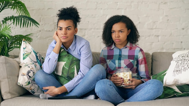 Zwei lockige Freundinnen gemischter Rassen sitzen auf der Couch und sehen sich zu Hause einen sehr gruseligen Film im Fernsehen an