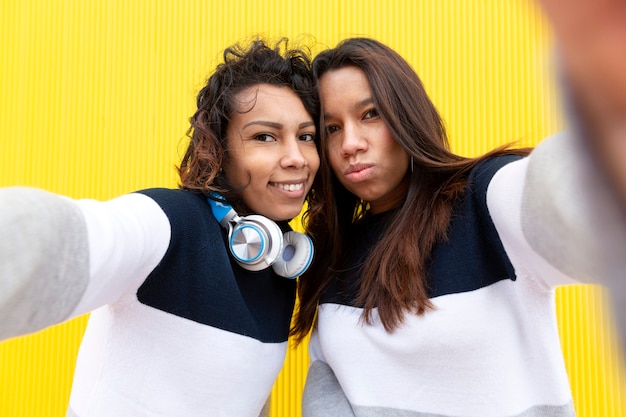 Zwei lustige hispanische Mädchen, die Selfie-Foto auf Handy nehmen. Sie sind auf gelbem Grund isoliert. Konzept der Freundschaft.