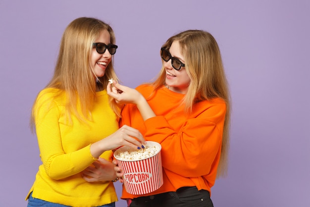 Zwei lustige junge blonde Zwillingsschwestern Mädchen in 3D-Imax-Brille, die Filmfilme ansehen und Popcorn isoliert auf pastellvioletter blauer Wand halten Menschen-Familien-Lifestyle-Konzept.
