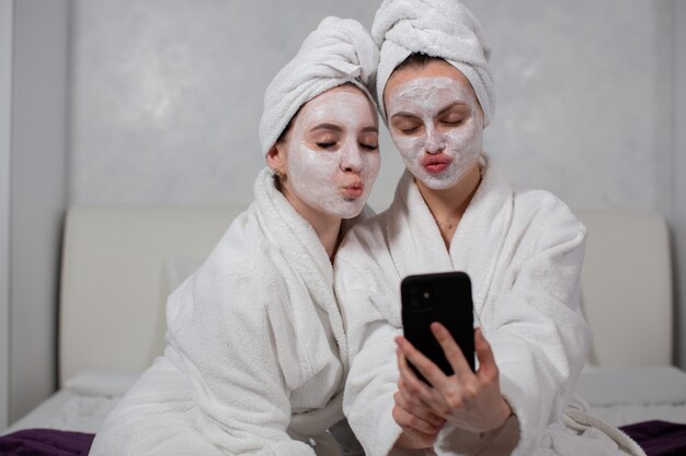 Zwei lustige Mädchenfreundinnen in Bademänteln, Gesichtsmasken und Handtüchern auf dem Kopf machen Selfies am Telefon. Foto in hoher Qualität