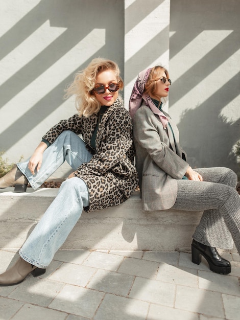 Zwei lustige, stylische, hübsche junge Freundinnen in modischer Herbstkleidung mit Sonnenbrille sitzen auf der Straße in der Nähe einer Betonwand im Sonnenlicht