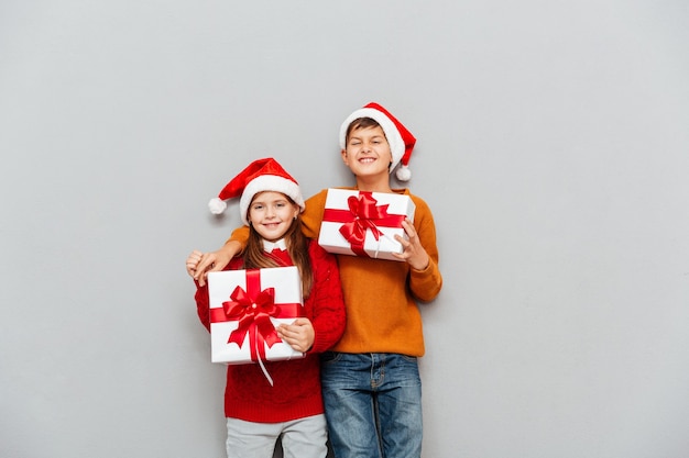 Zwei lustige süße Kinder in Weihnachtsmannmützen mit Geschenkboxen stehen zusammen