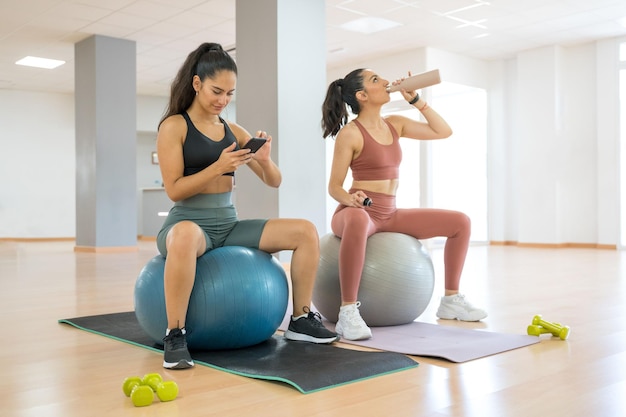 ZWEI MÄDCHEN MACHEN PILATES UND YOGA, WÄHREND EINS TELEFONIERT UND EIN ANDERES WASSER TRINKT