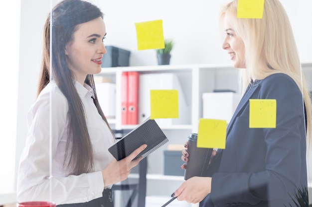 Zwei Mädchen, die im Büro sprechen. Mädchen sind ein Dialog in der Nähe einer transparenten Tafel mit Aufklebern.