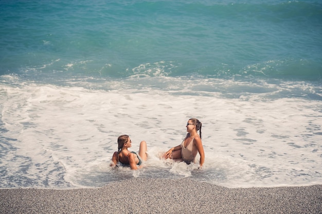 Zwei Mädchen, Freundin, sitzen am Sandstrand des Meeres und die Wellen haben sie an einem sonnigen, warmen Tag in Badeanzüge getränkt