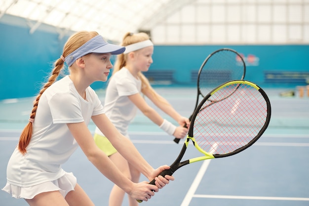 Zwei Mädchen im Teenageralter in Activewear stehen im Stadion mit gebeugten Beinen in den Knien und halten Tennisschläger bereit, um den Ball während des Spiels zu schieben