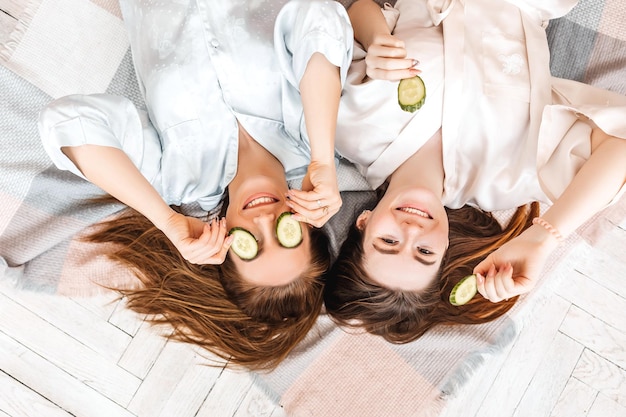 Zwei Mädchen machen hausgemachte Schönheitsmasken für Gesicht und Haare Gurken für die Frische der Haut um die Augen Frauen kümmern sich um jugendliche Haut Freundinnen lachen zu Hause auf Kissen auf dem Boden liegend