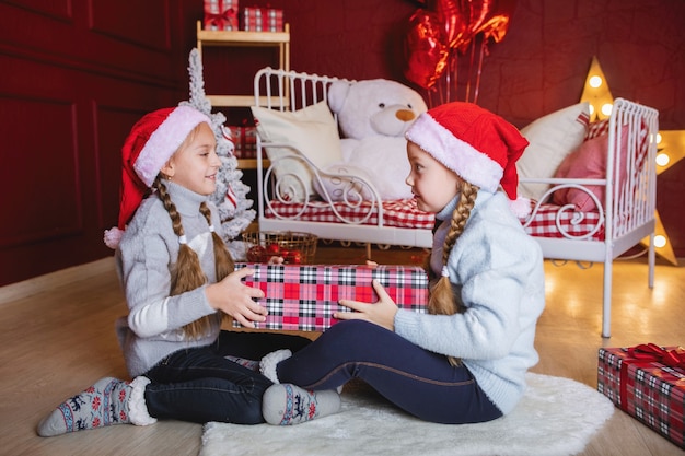 Zwei Mädchen mit Zöpfen in roten Weihnachtsmützen mit Geschenkboxen sitzen auf dem Boden neben dem Bett.