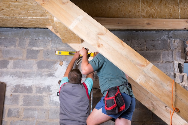 Zwei Männer arbeiten zusammen, um einen Holzrahmen für Treppen zu bauen, die in den Keller eines unfertigen Hauses führen, und messen die Ebenheit der Stufen mit dem gelben Nivellierwerkzeug