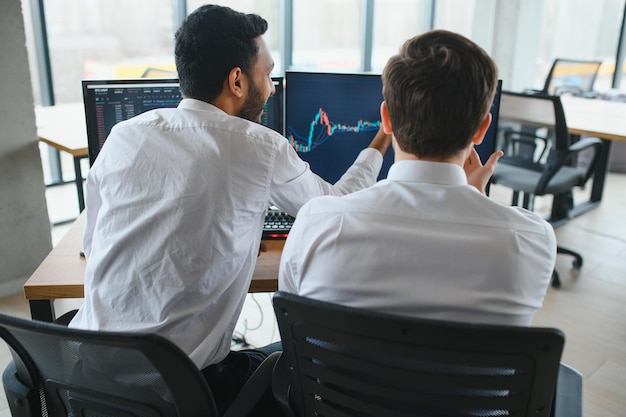 Zwei Männer, die am Schreibtisch im Büro sitzen, überwachen gemeinsam Aktiendaten-Kerzendiagramme auf dem Bildschirm und analysieren den Preisfluss, der fröhlich lächelt und ein Gewinn-Teamwork-Konzept hat