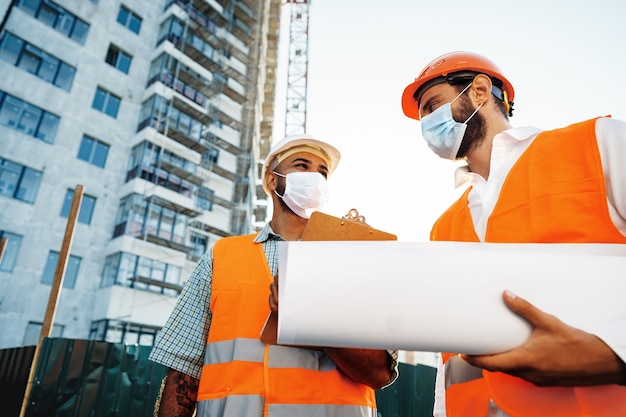 Zwei Männer in Arbeitskleidung und medizinischen Masken, die mit Blaupausen auf Objekten arbeiten