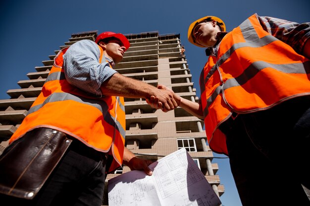 Zwei Männer in Hemden, orangefarbenen Arbeitswesten und Helmen schütteln sich vor dem Hintergrund eines mehrstöckigen Gebäudes die Hand.