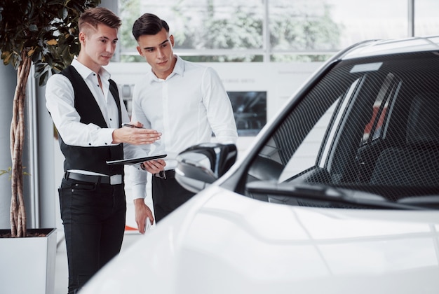 Zwei Männer stehen im Ausstellungsraum gegen Autos. Nahaufnahme eines Verkaufsleiters in einem Anzug, der ein Auto an einen Kunden verkauft. Der Verkäufer gibt dem Kunden den Schlüssel.