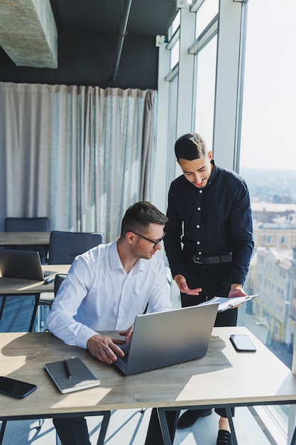 Zwei männliche Kollegen diskutieren Geschäftsprojekte und schauen während eines Meetings in einem modernen Besprechungsraum mit Glaswänden auf den Laptop-Bildschirm